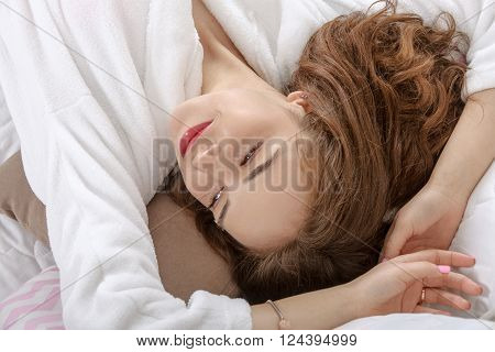 Girl woke up in a white bed in the morning. She is dressed in a white robe, her eyes closed and she lies on her back throwing her hands behind her head. Concept: lifestyle, health and beauty.