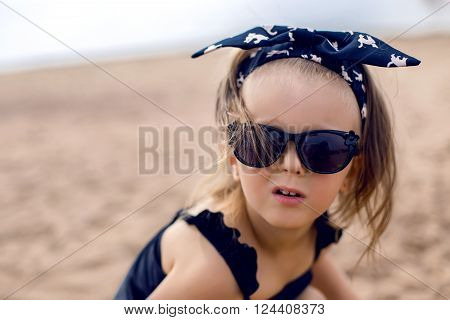 a little girl stands on the shore of the beach in a black bathing suit and black glasses