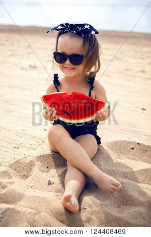 a little girl stands on the shore of the beach in a black bathing suit and black glasses eating juicy watermelon sitting