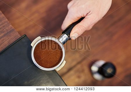 Overhead shot of a person's hand holding the handle of shiny new portafilter for an espresso machine containing a perfectly level amount of fresh ground coffee, with a tamper visible on counter below