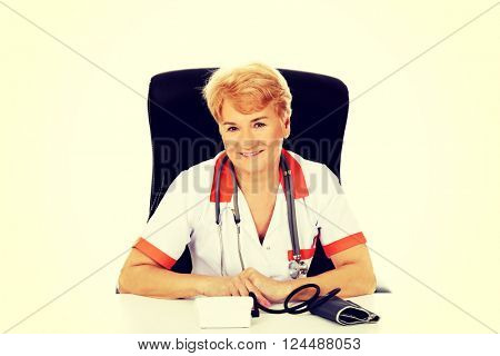 Smile elderly female doctor or nurse sitting behind the desk with bloog preasure gauge