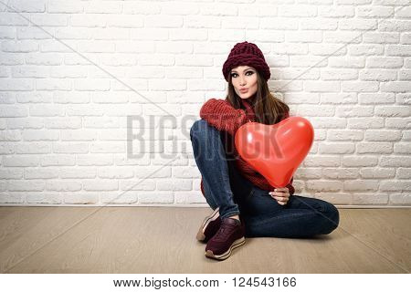 Portrait of attractive young woman sitting near white bricks wall and holding red heart balloon.