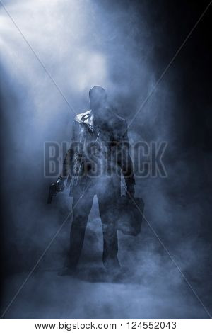 Faceless man holding a gun and a briefcase standing in haze.