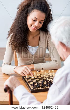 Young pretty happy carer plying chess with her older ward