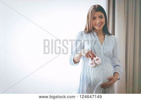 Pregnant woman standing near window with baby's bootees in the room