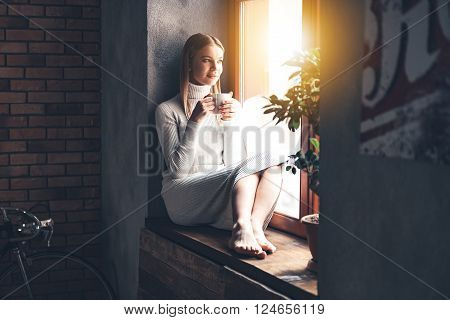 Making plans for new day. Beautiful young woman holding coffee cup and looking through window while sitting at windowsill at home