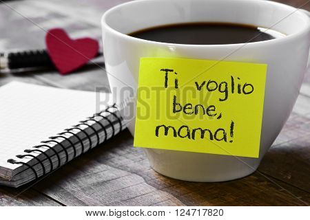 the sentence ti voglio benne mama, I love you mom written in italian in a yellow sticky note placed in a cup of coffee, on a wooden table next to a notepad and a red heart in the background