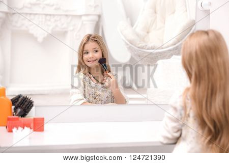 Little beautiful girl applying makeup in front of mirror. Nice cozy white bedroom. Girl using brush for makeup
