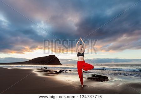 Summer sunrise yoga session on beautiful Playa de La Tejita beach with view on atlantic ocean and Punta Roja rock - tropical Tenerife island Canary in Spain. Vriksha-asana tree pose