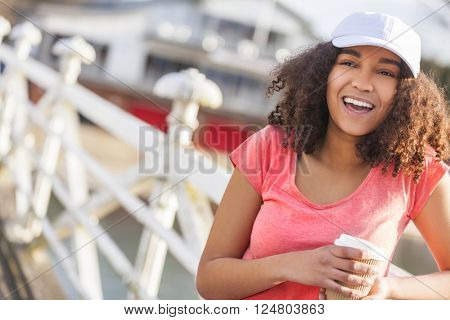 Beautiful happy mixed race African American girl teenager female young woman with perfect teeth smiling drinking takeaway coffee outside wearing white baseball cap and pink t-shirt