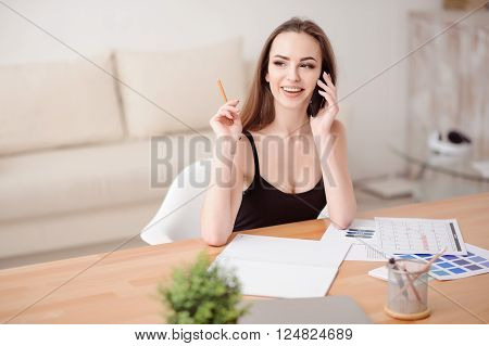 Nice interlocutor. Cheerful content positive beautiful girl sitting at the table and talking on mobile phone while being involved in work