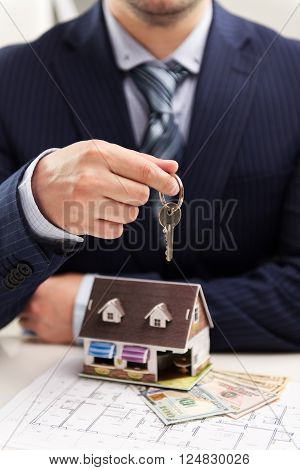 Young businessman broker offering house for rent holding keyring. Closeup of male real estate agent sitting in the office with building model on desk.