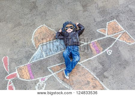 Funny little kid boy flying by a plane picture painting with colorful chalk. Creative leisure for children outdoors in summer.