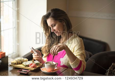Young happy woman eating sushi for lunch at a small cafe and a healthy lifestyle, working on a smartphone. Smiling, fashionable, in a yellow sweater.
