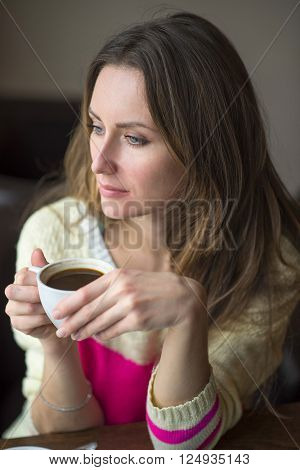 Young happy woman holding a cup of hot coffee or tea, sitting in a cafe looking out the window, lifestyle, breakfast or dinner to spend time relaxing, healthy lifestyle, fashion, in a yellow sweater.