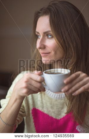 Young happy woman holding a cup of hot coffee or tea, sitting in a cafe looking out the window, lifestyle, breakfast or dinner to spend time relaxing, healthy lifestyle, fashion, in a yellow sweater.