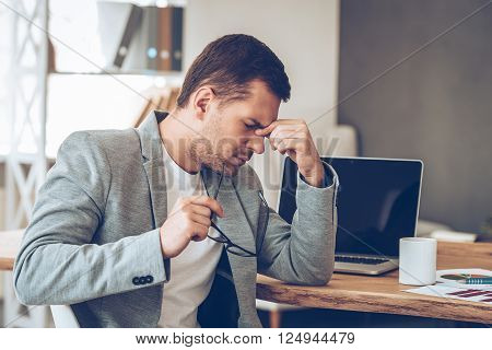 Feeling tired. Frustrated young handsome man looking exhausted while sitting at his working place and carrying his glasses in hand