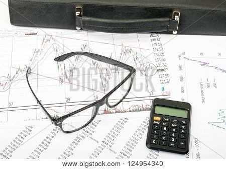 Glasses and suitcase with calculator on grpahical charts background, business concept