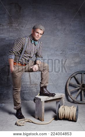 Handsome guy in checkered shirt with blue bow tie posing near gre concrete wall with wooden wheel.