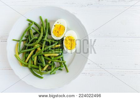 Cooked green beans with boiled eggs in white plate on wooden background with space for text. Healthy vegetarian food concept. Top view. Copy space.