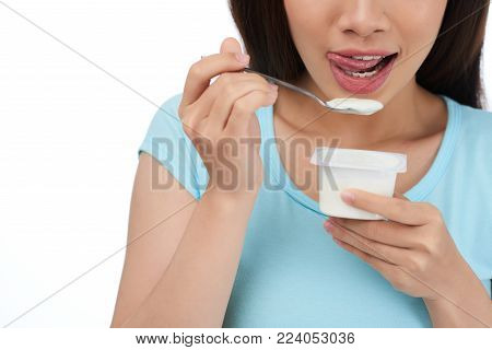 Close-up shot of unrecognizable dark-haired woman licking her lips while holding yogurt cup and spoon in hands, isolated on white background