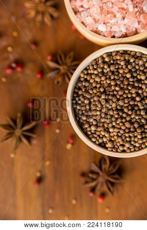Pink Himalayan salt and coriander seeds in white bowls on wooden surface covered with anise and red peppercorn