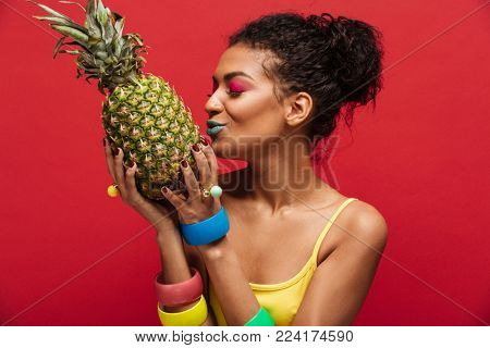 Mixed-race woman with fashion makeup in yellow shirt having detox kissing fresh ripe pineapple holding in hands isolated over red background
