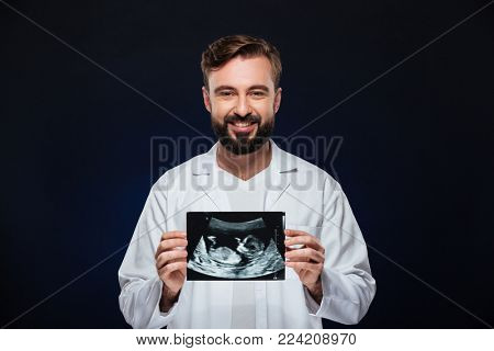 Portrait of a friendly male doctor dressed in uniform showing baby x-ray isolated over dark background