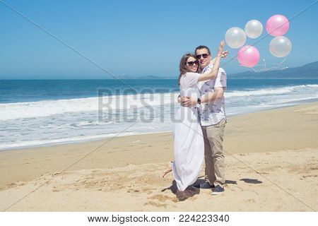 Young romantic couple in white spending vacation or weekend on the beach, celebrating with pink balloons, hugging, for header webside
