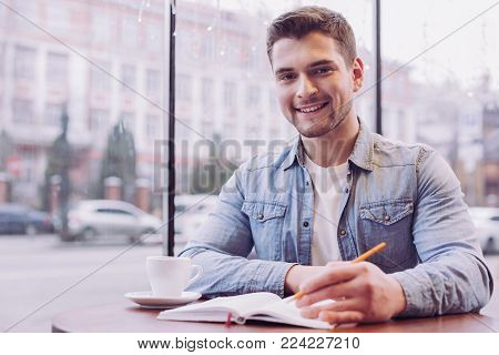 Self education. Bearded young male student writing down information while smiling and looking at the camera