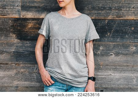 Summer day. Front view. Young millennial woman dressed in gray t-shirt is stands against gray wood wall. Mock up. Space for logo, text, image.