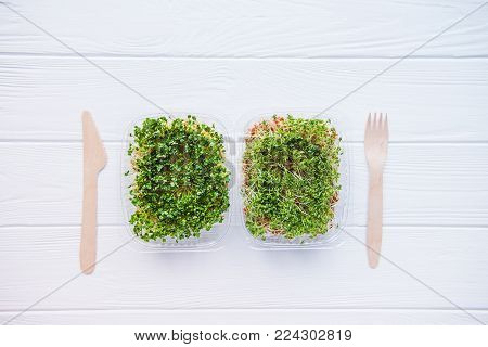 Top view plastic boxes with fresh organic sprout micro greens and wooden cutlery on the white table. Healthy Raw diet food concept. Copy space for text. Selective focus
