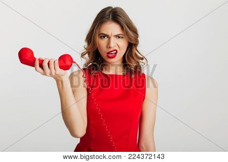 Portrait of a confudsed young woman dressed in red dress holding telephone isolated over white background