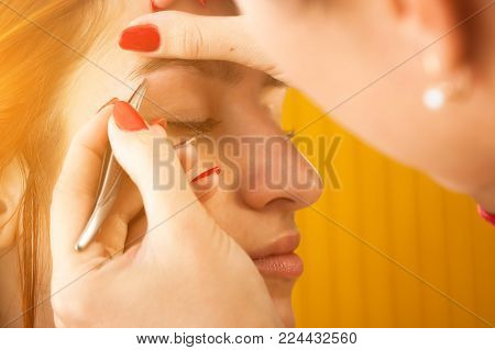 Close up of the master on the eyebrows corrects the shape of the roach with tweezers  red-haired beautiful woman on a white background; view from above