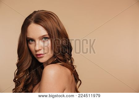 Beauty portrait of mystery ginger woman with long hair posing sideways and looking at the camera over cream background