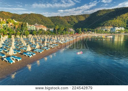 Aerial view of beautiful beach with chaise-lounges and swimming woman on the air mattress in the sea against mountains at sunset. Summer seascape with girl, blue water, sky. Top view. Icmeler, Turkey