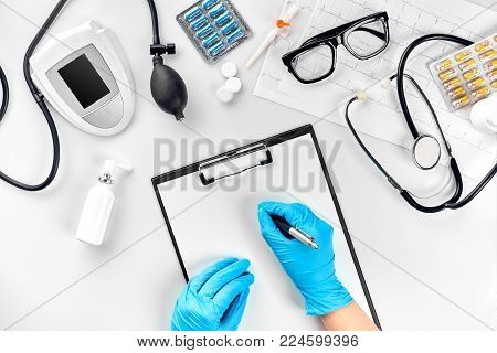 Doctor s table, tools, medical instruments, therapist tonometer, blood pressure, work in hospital on white background flat lay. The doctor's hand in a blue glove records the indicators on a white paper blank. Top view. Still life. Copy space