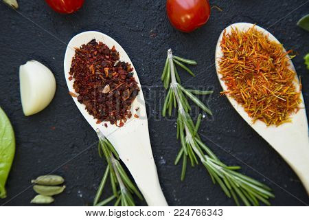 Cooking a hot spicy meal with a set of spices and herbs in wooden spoons, fresh rosemary, lettuce, garlc, and cayenne chili pepper, black concrete background, top view