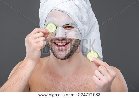 cheerful bearded man with collagen mask and cucumber slices, isolated on grey, skin care concept