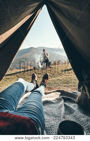 Panoramic view. Woman enjoying the view of mountain range from the tent while her boyfriend resting near the campfire