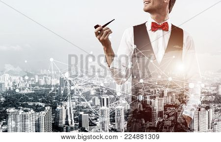 Double exposure of confident and young businessman in smart-casual wear smoking pipe and modern cityscape with business and media interface. Concept networking for modern business.
