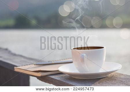 cup of hot coffee for brackfast and note book with pen on wooden balcony near river and mountain background