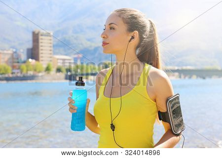 Thirsty Female Athlete With Armband For Smart Phone Holding Power Drink And Looking Away Outdoors.