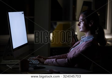 Young Businesswoman Sitting At Her Workplace In Front Of Computer Monitor And Typing In Dark Office 