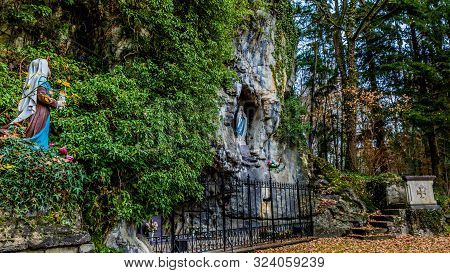 Statue Of Bernadette Praying To Our Lady Of Lourdes In A Natural Grotto With A Metal Fence On A Magi
