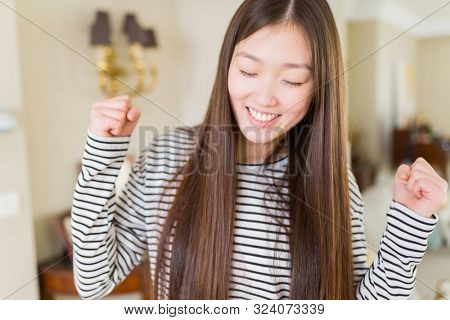 Beautiful Asian woman wearing stripes sweater over living room background excited for success with arms raised celebrating victory smiling. Winner concept.