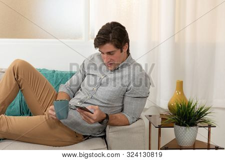 Front view close up of a happy young Caucasian man holding a cip of coffee and using a smartphone reclining on the sofa in his sitting room