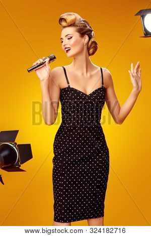 Beautiful young woman sings on stage with a microphone in the light flashes. Pin-up style in clothes, hair and make-up.