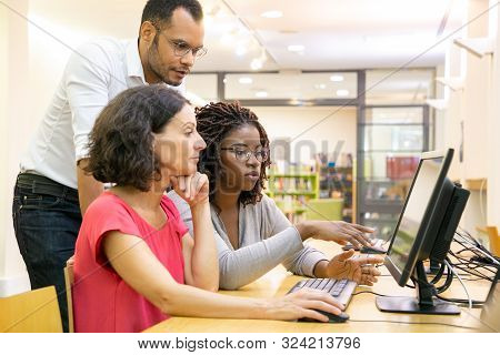 Tutor Helping Students In Computer Class. Man And Women Sitting And Standing At Desk, Using Desktop,
