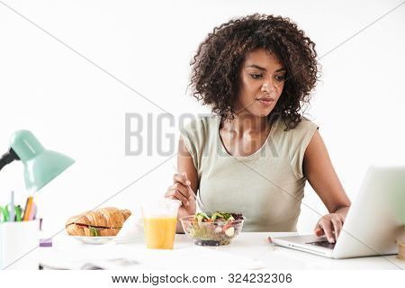 Beautiful young african woman sitting at the desk isolated over white background, having lunch while working on laptop computer